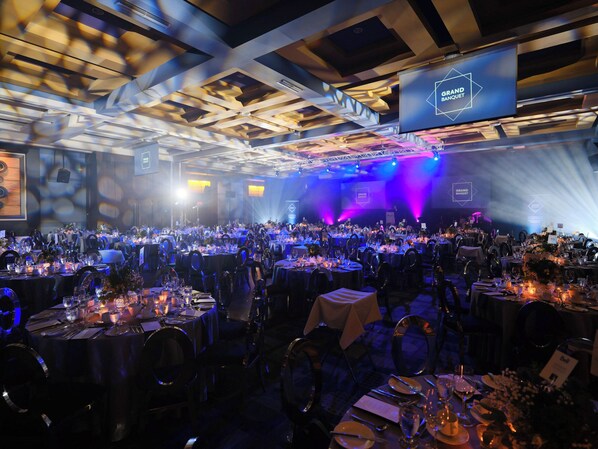 A banquet hall with tables set for a formal event, featuring a high ceiling with geometric patterns and a stage with lighting.
