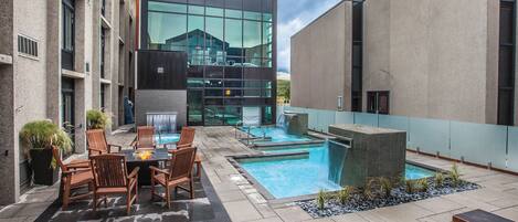 A rooftop pool area with a glass-enclosed building, outdoor seating, and a waterfall feature.