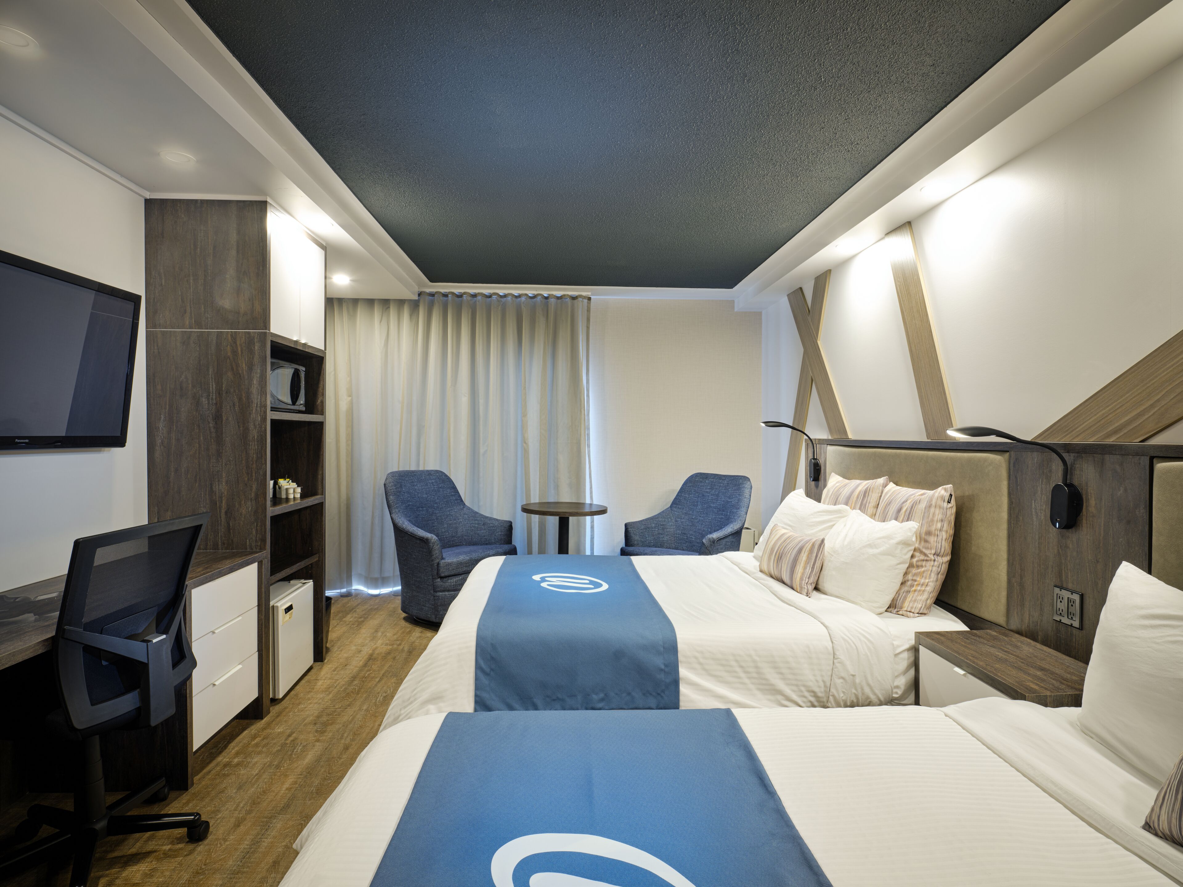 A hotel room with a bed, two chairs, a desk, a TV, and a small table.