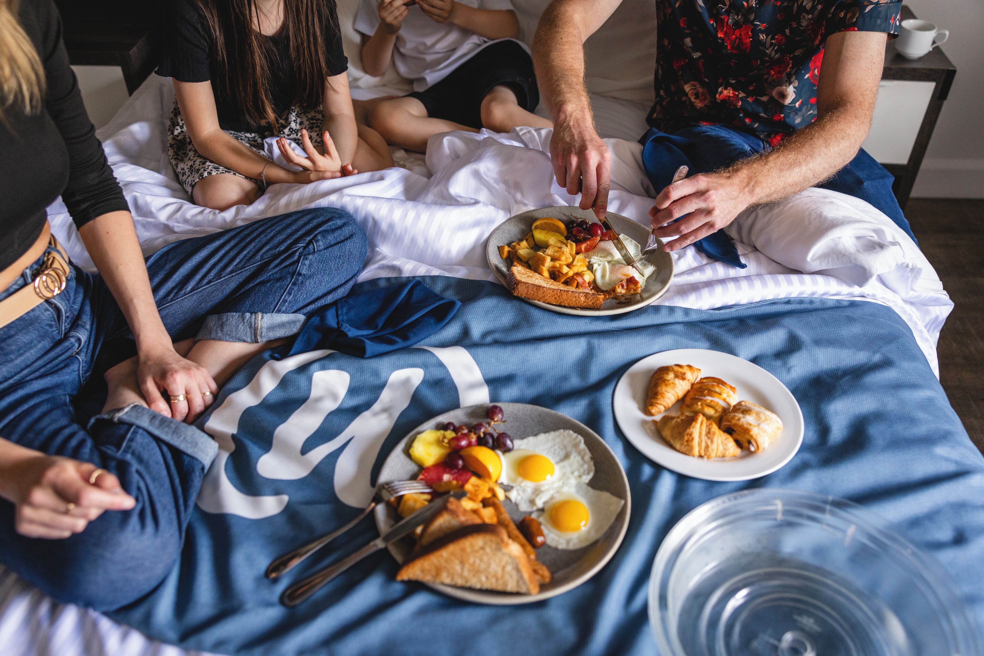 Een groep mensen zittend op een bed met een blauwe deken, die ontbijt eten.