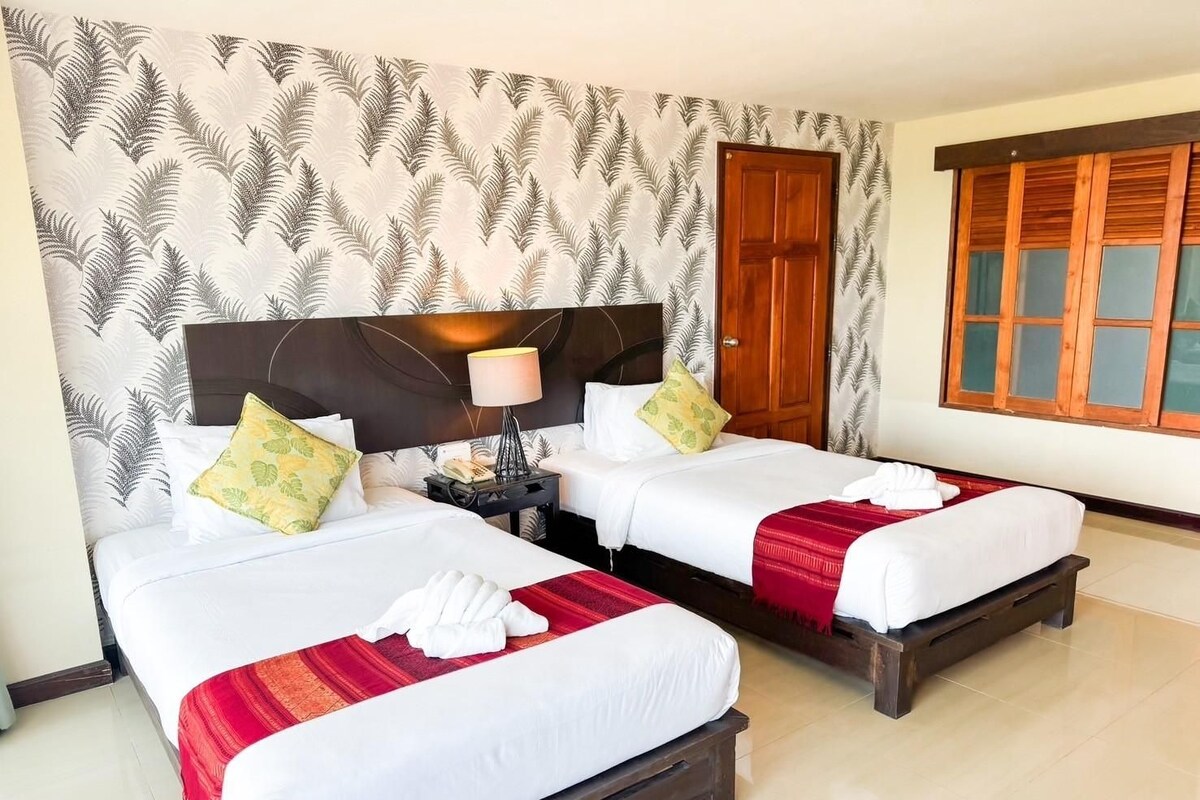 A hotel room with two beds, a wooden headboard, a nightstand, a lamp, and a window with wooden shutters.