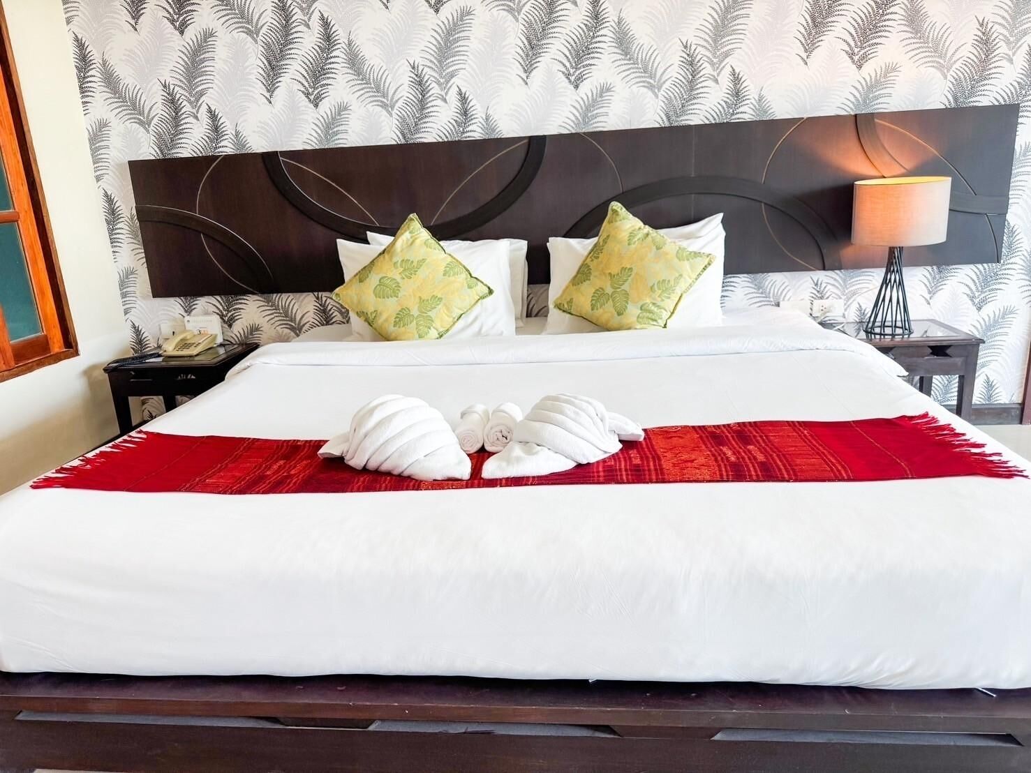 Una cama bien tendida con sábanas blancas, dos almohadas estampadas con hojas verdes y una manta roja.