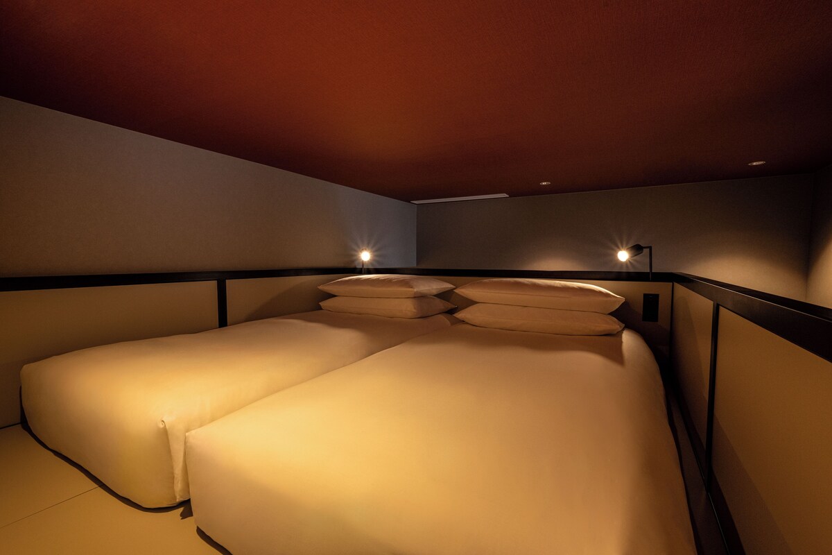 A hotel room with two beds, a nightstand, and a wall-mounted lamp.