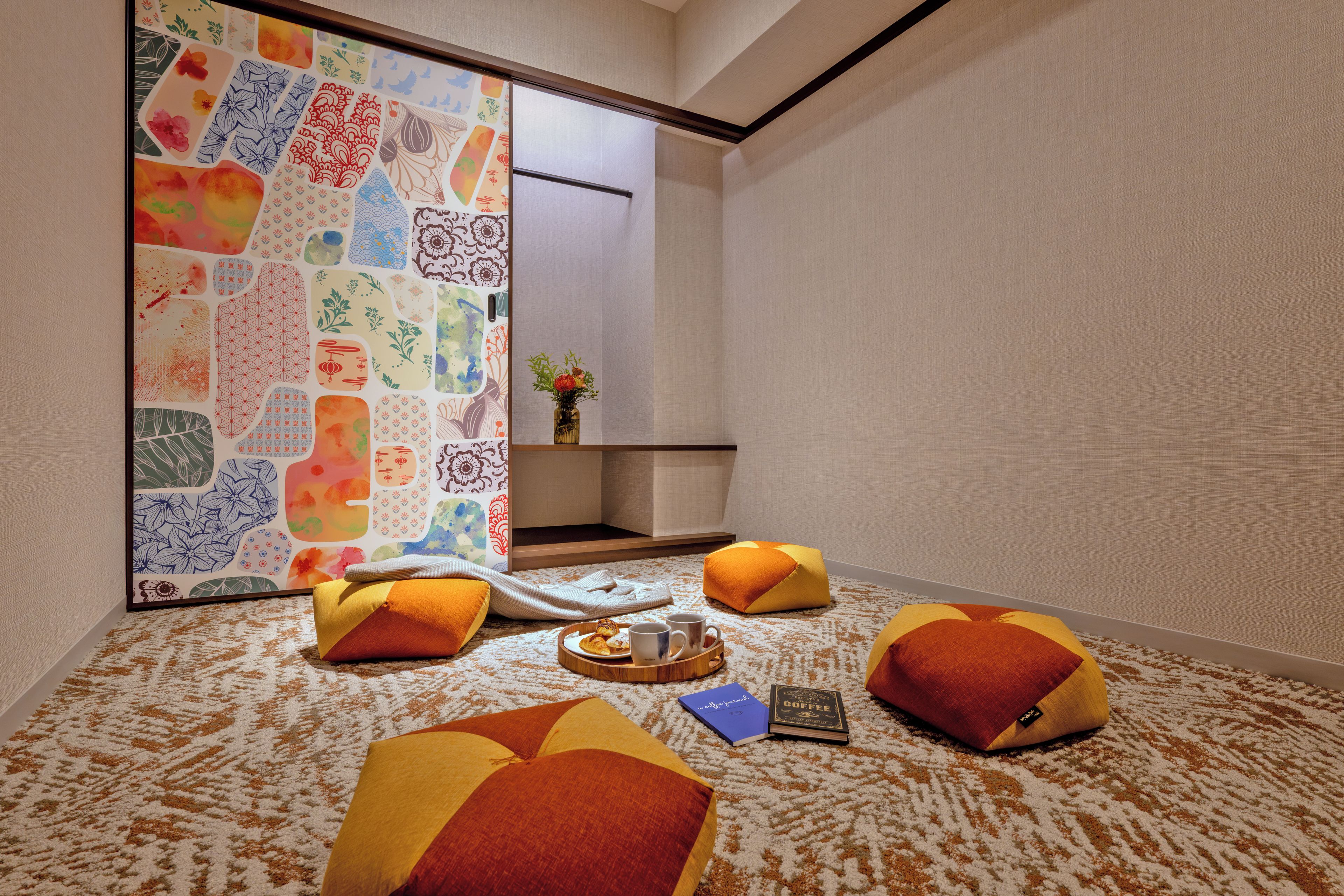 Una habitación con alfombra estampada, una puerta corredera con obra de arte colorida y una pequeña mesa con un libro y una taza.