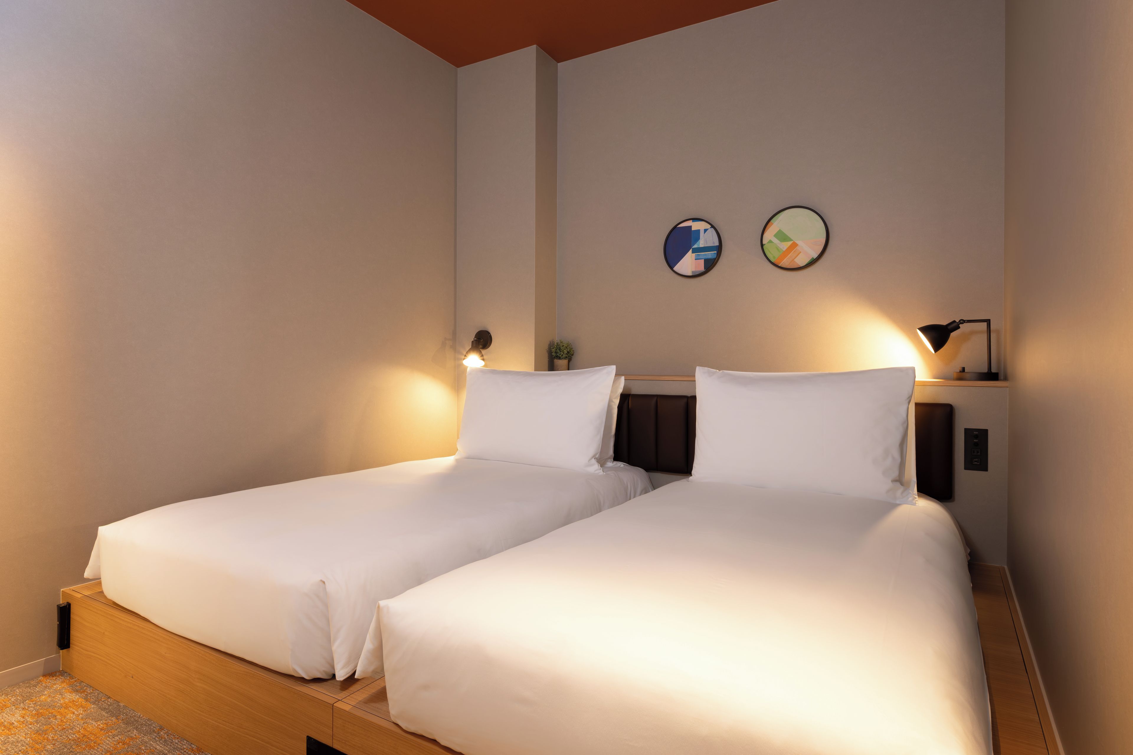 Quarto de hotel com duas camas de solteiro, uma pequena mesa de cabeceira com abajur e uma obra de arte na parede.