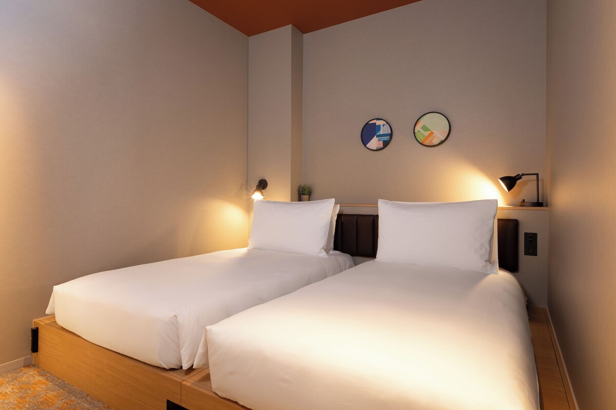 A hotel room with two single beds, a small bedside table with a lamp, and a wall-mounted artwork.