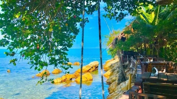 A coastal view with clear blue water, rocky outcrops, and a wooden deck with seating. - Haadson Resort (Ko Pha-ngan)