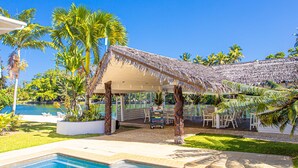 A pool area with a thatched-roof structure, palm trees, and a clear blue sky.