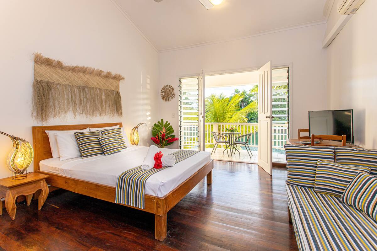 A spacious bedroom with a wooden bed, a sofa, and a balcony with a view of greenery.