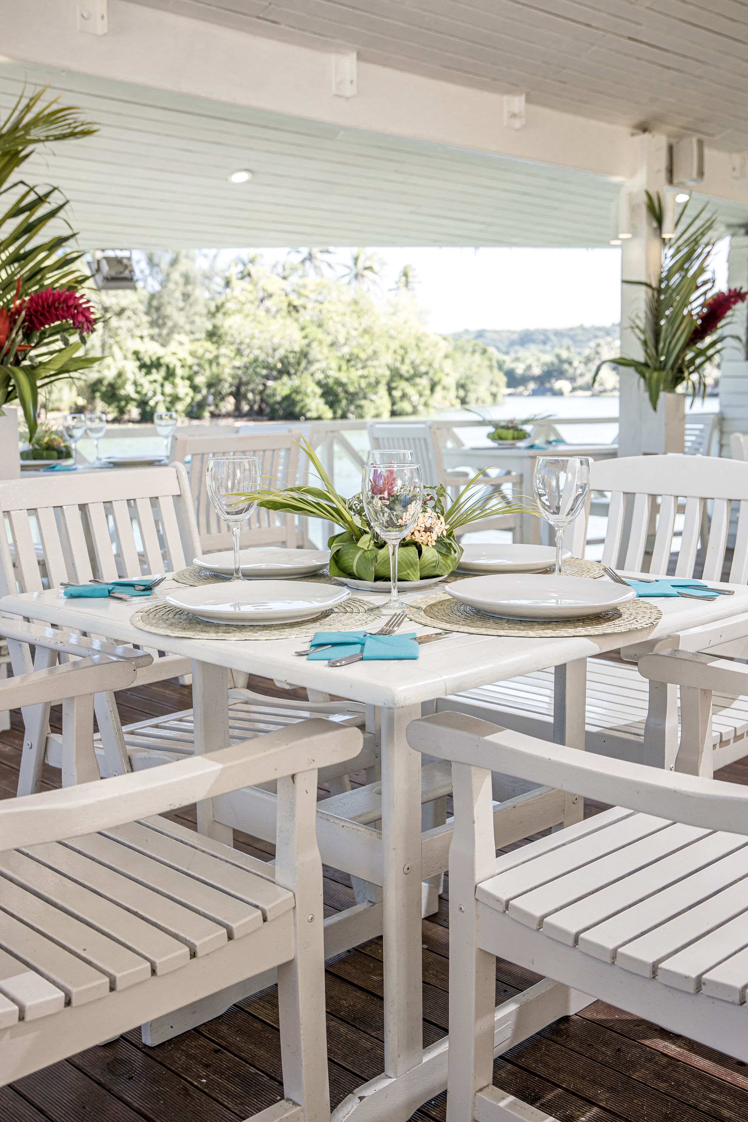 A white outdoor dining area with tables and chairs, surrounded by greenery and a view of the landscape.