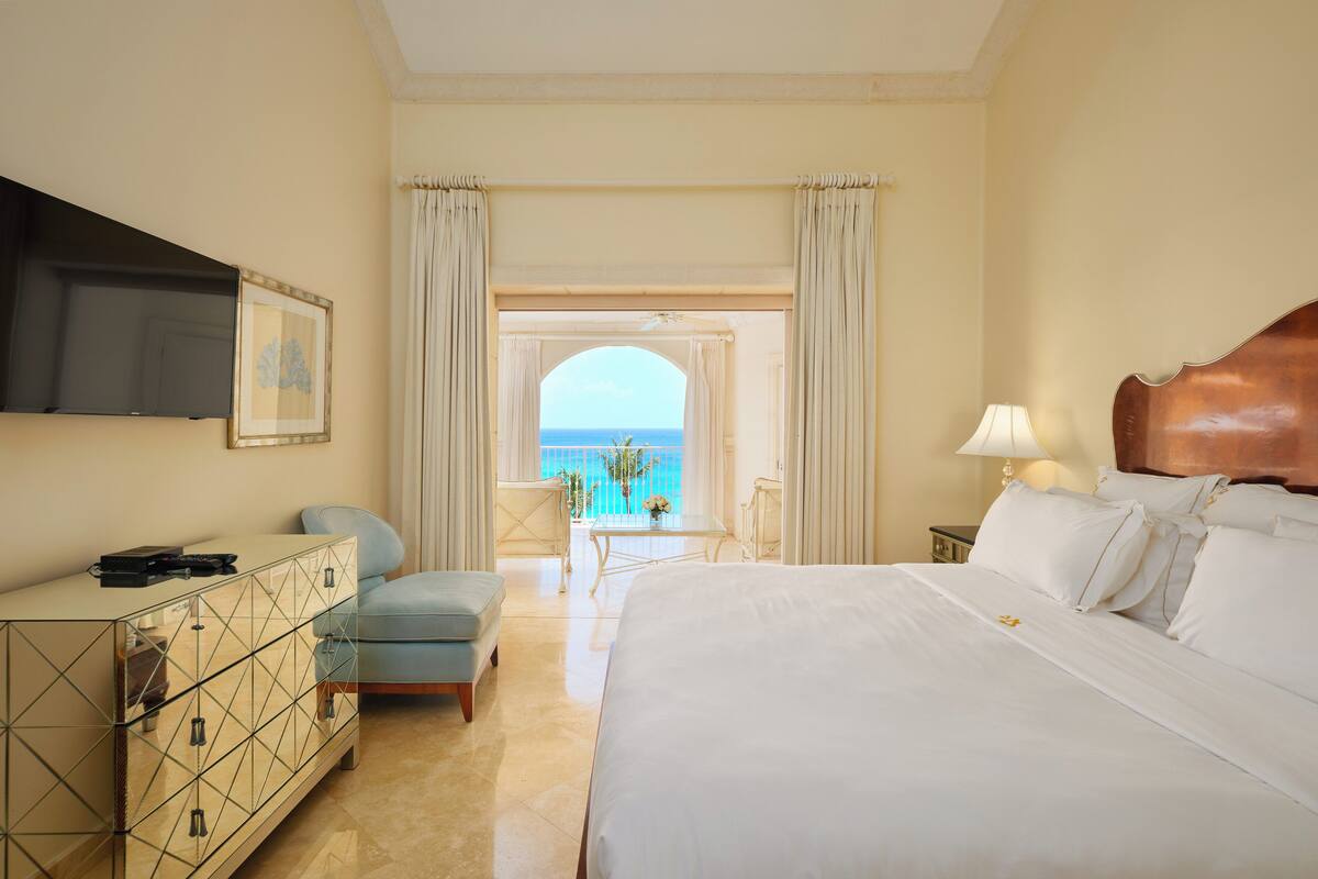 A hotel room with a large bed, a TV mounted on the wall, a desk with a chair, and a view of the sea through an arched doorway.