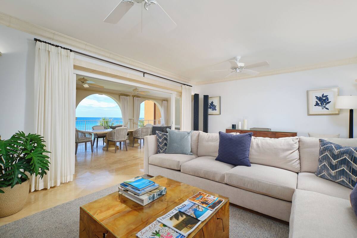 A spacious living room with a beige sofa, a wooden coffee table with magazines, a large potted plant, and a view of the ocean through an arched doorway.