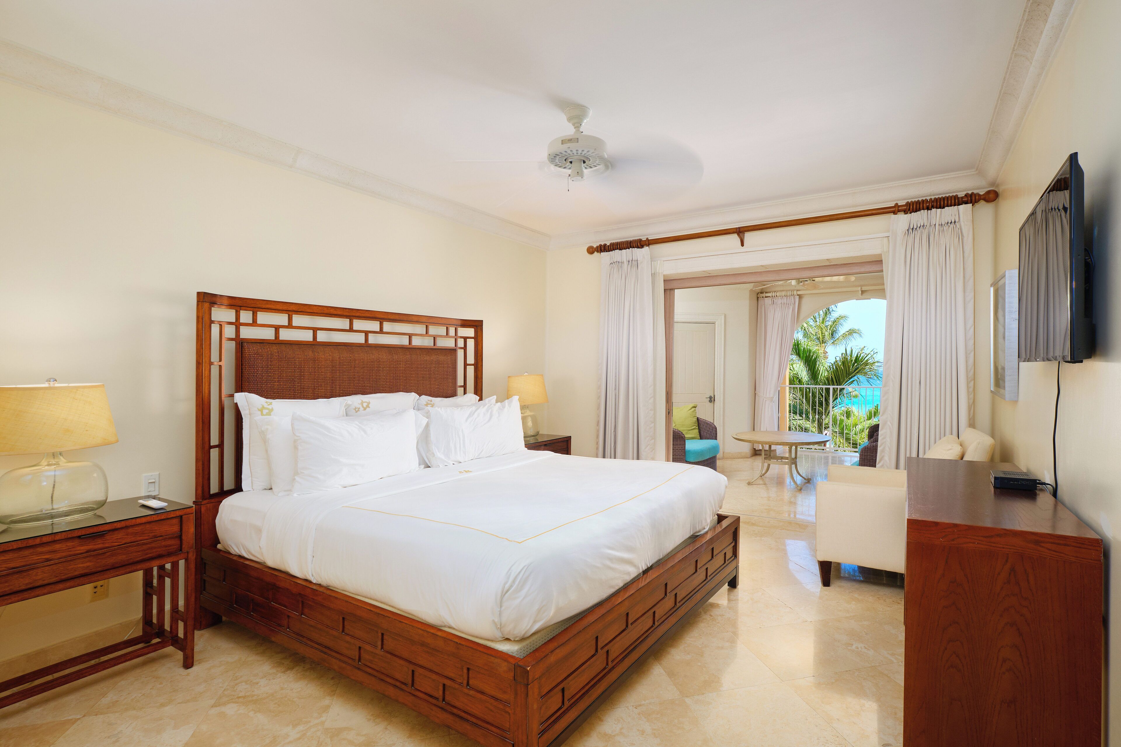 A hotel room with a large bed, wooden headboard, two bedside tables with lamps, a TV, and a balcony with curtains.