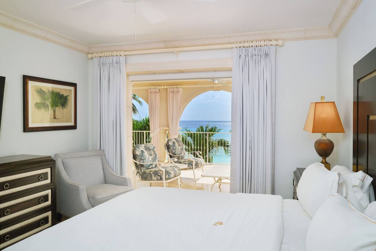 A hotel room with a bed, a chair, a dresser, a lamp, and a view of the ocean through a balcony.