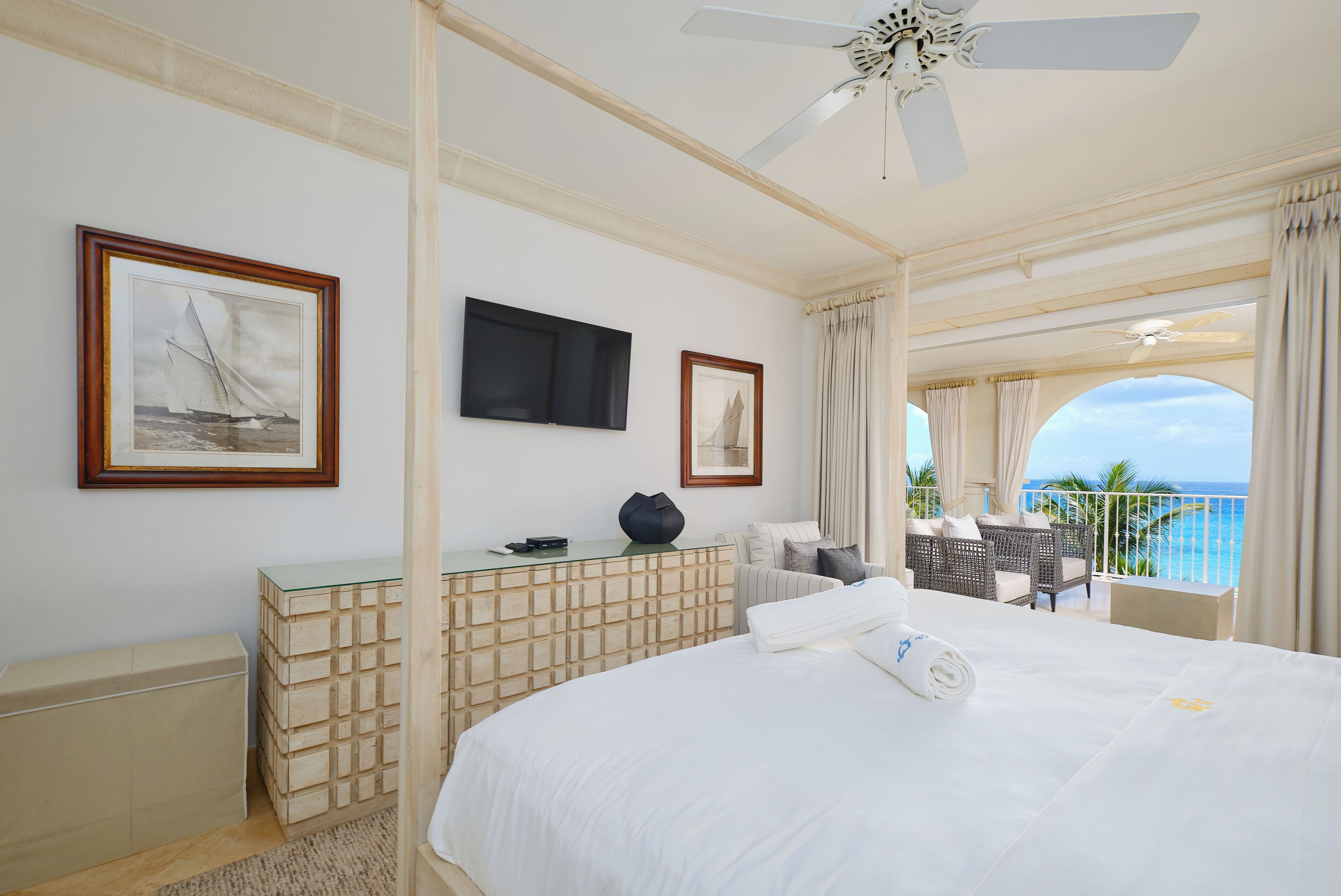 A hotel room with a large bed, a flat-screen TV, a desk with a glass top, and a balcony with a view of the ocean.