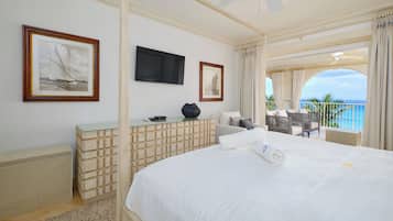 A hotel room with a large bed, a flat-screen TV, a desk with a glass top, and a balcony with a view of the ocean.