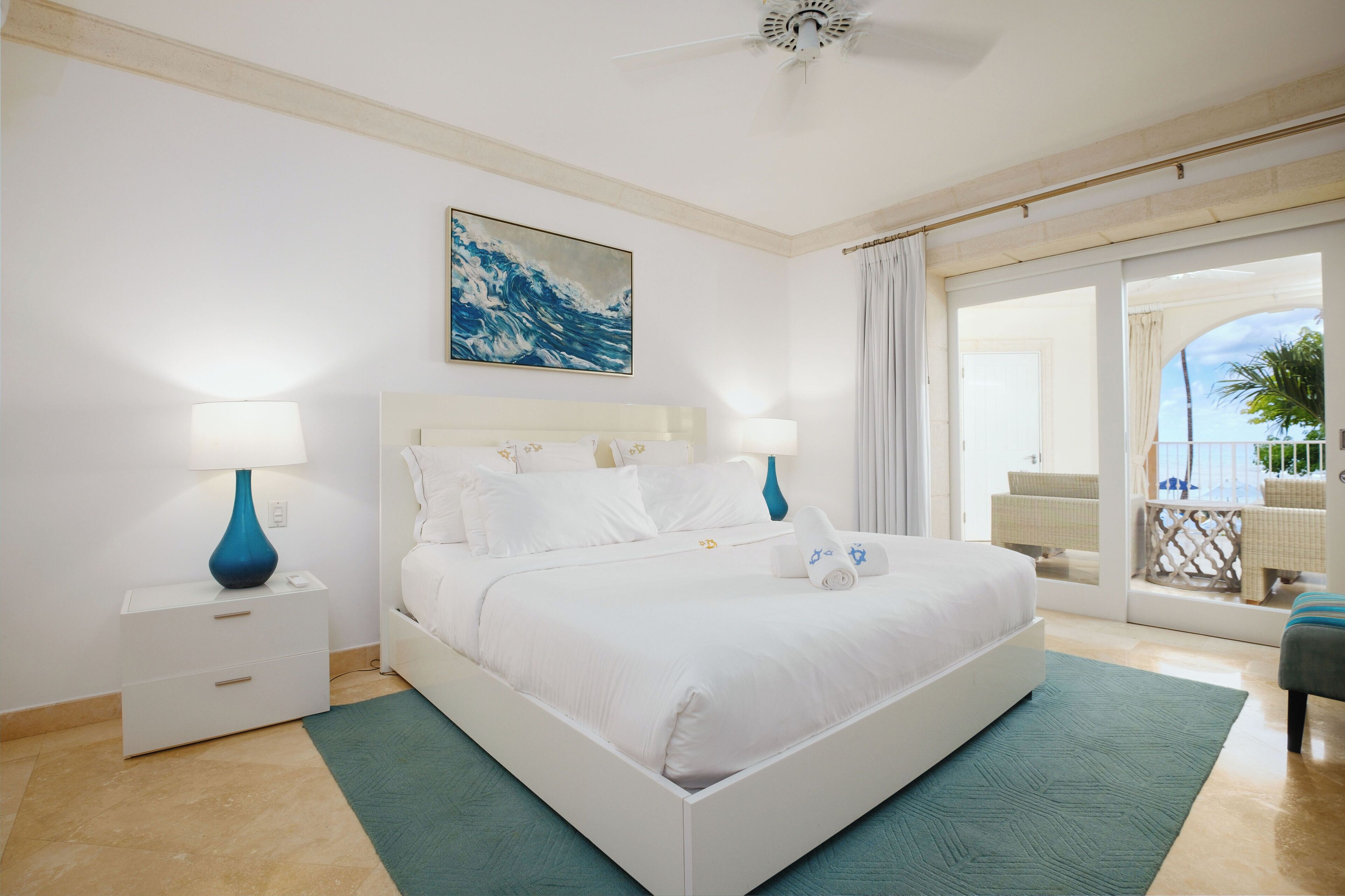 A bedroom with a bed, nightstands, a ceiling fan, a painting, and a view of the outside balcony.