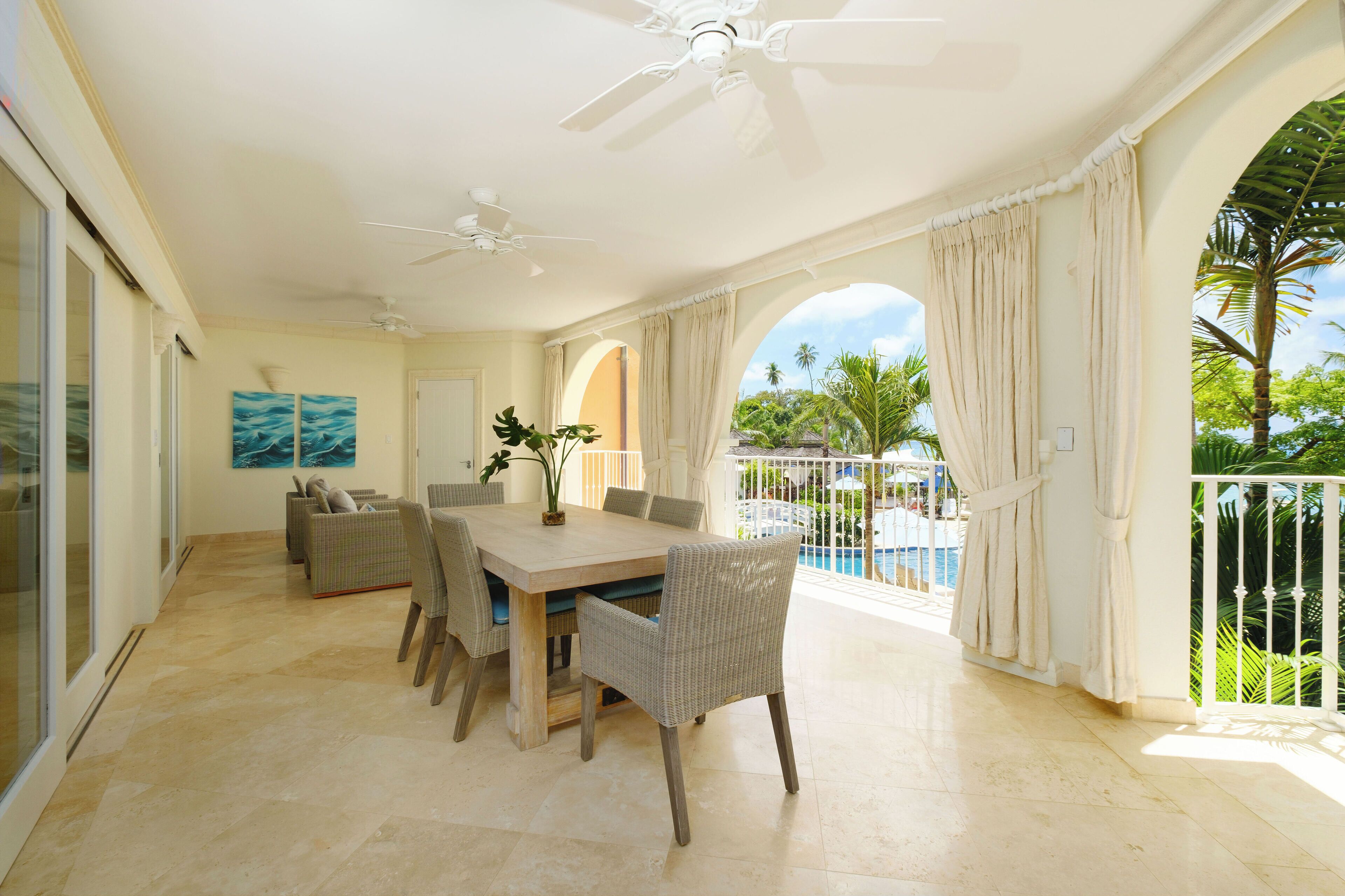 A spacious dining area with a large table and chairs, a ceiling fan, and a view of a pool and palm trees.