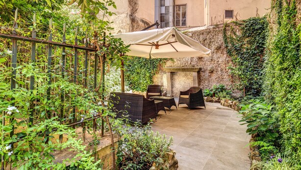 Un patio exterior con mobiliario de mimbre y un gran paraguas, rodeado de abundante vegetación y un muro de piedra.