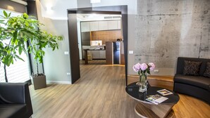 Una zona de recepción moderna con suelo de madera, un sofá negro, una mesa de centro de vidrio con un jarrón de flores y vistas a una cocina.