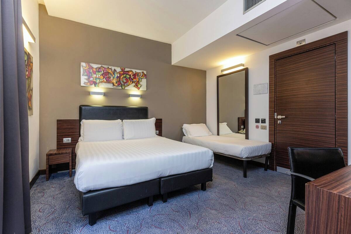 A hotel room with a large bed, a smaller bed, a desk, a chair, a mirror, and a wall with a colorful artwork.