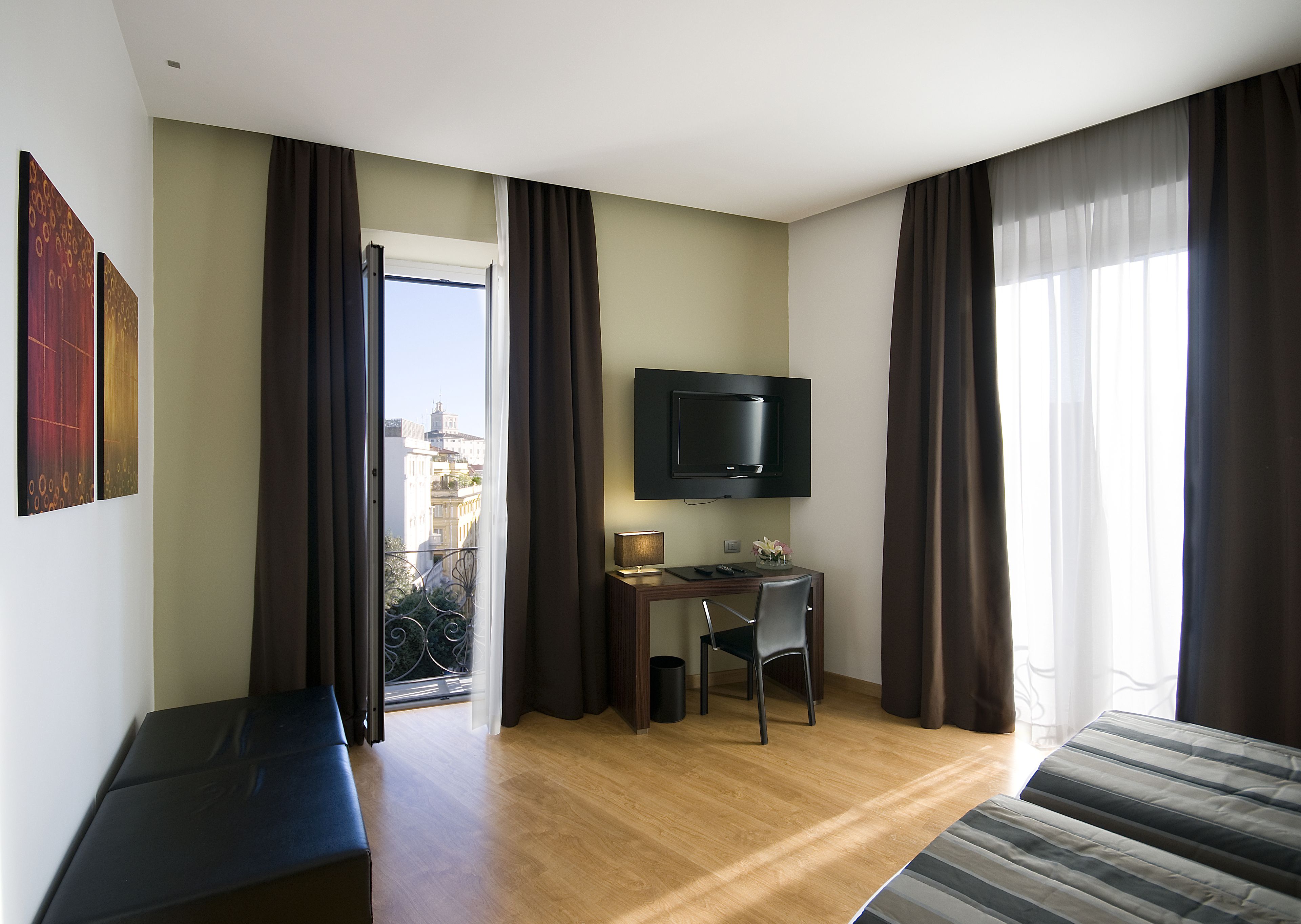 Una habitación de hotel con cama, un escritorio con silla, un televisor y un balcón con cortinas.