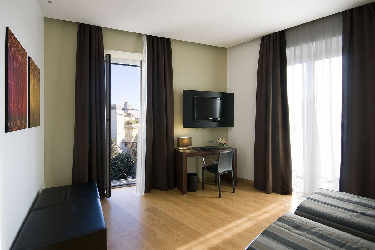 A hotel room with a bed, a desk with a chair, a television, and a balcony with curtains.