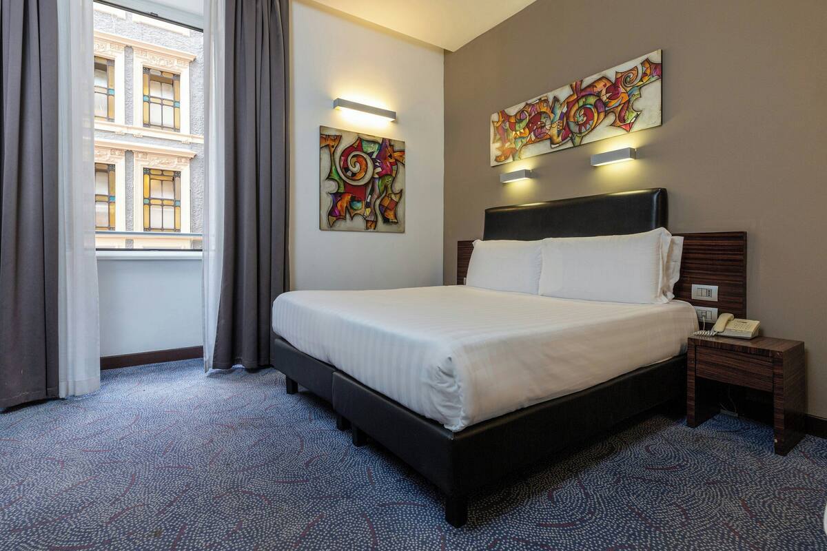 A hotel room with a bed, a nightstand with a phone, a window with curtains, and two wall-mounted light fixtures.