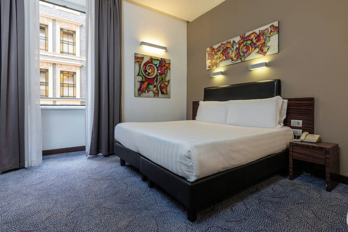 A hotel room with a bed, a nightstand with a phone, a window with curtains, and two wall-mounted light fixtures.