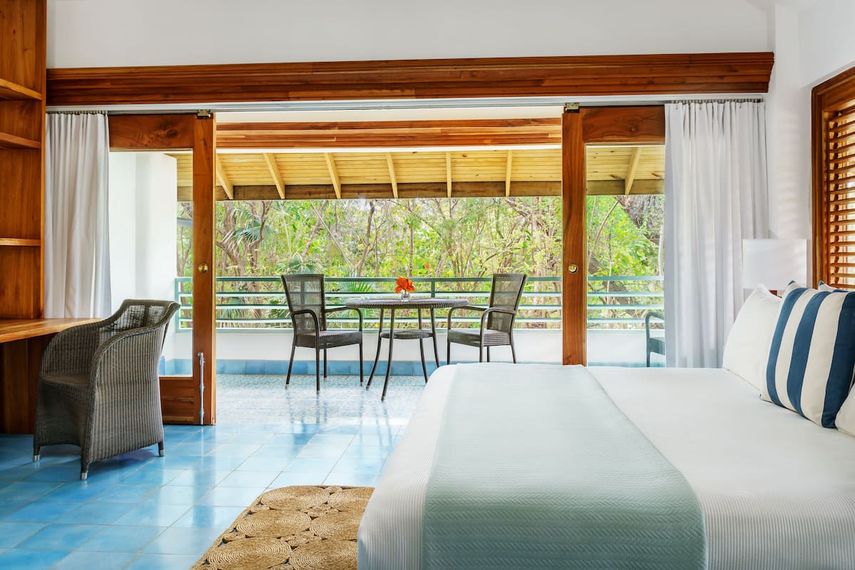 A bedroom with a bed, a desk, and a table with chairs. There is a view of greenery outside.