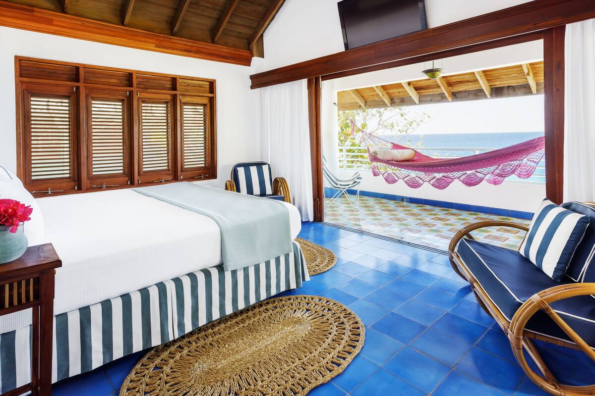 A bedroom with a bed, a wooden headboard, a wicker chair, a hammock, and a view of the ocean.