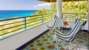 A balcony with a view of the ocean, a striped chair, and a small table with a flower.