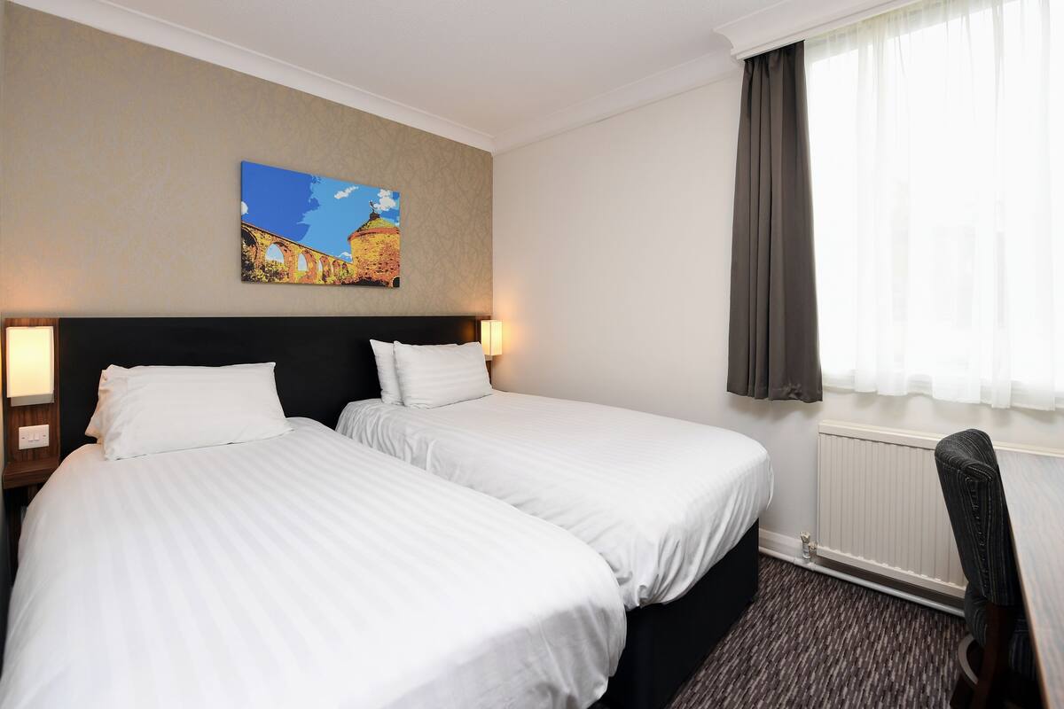 A hotel room with two beds, a painting of a bridge, a window with curtains, and a chair.
