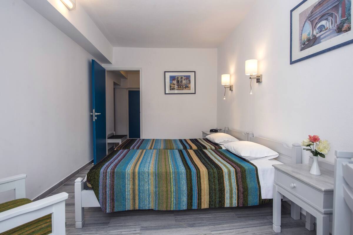 A hotel room with a bed, bedside table, and a nightstand. There is a door leading to another room and a framed picture on the wall.