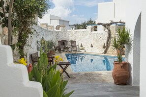 A pool area with stone walls, a wooden table, and chairs. - Aegeon Hotel (Paros)