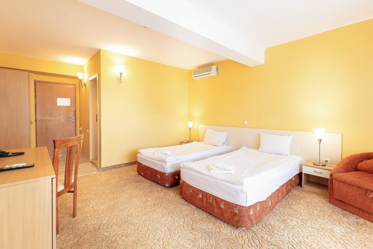 A hotel room with two single beds, a desk with a chair, a small table with a lamp, and a couch.