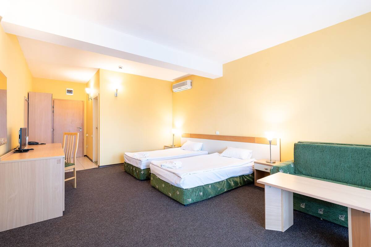 A hotel room with two single beds, a desk, a chair, a bench, and a green couch.