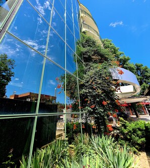 A modern building with reflective glass windows, surrounded by lush greenery and red flowers. - Juli (Sunny Beach)