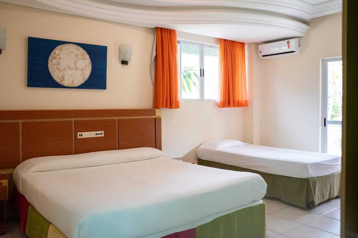Habitación de hotel con dos camas, un reloj de pared grande, un equipo de aire acondicionado y una ventana con cortinas naranjas.