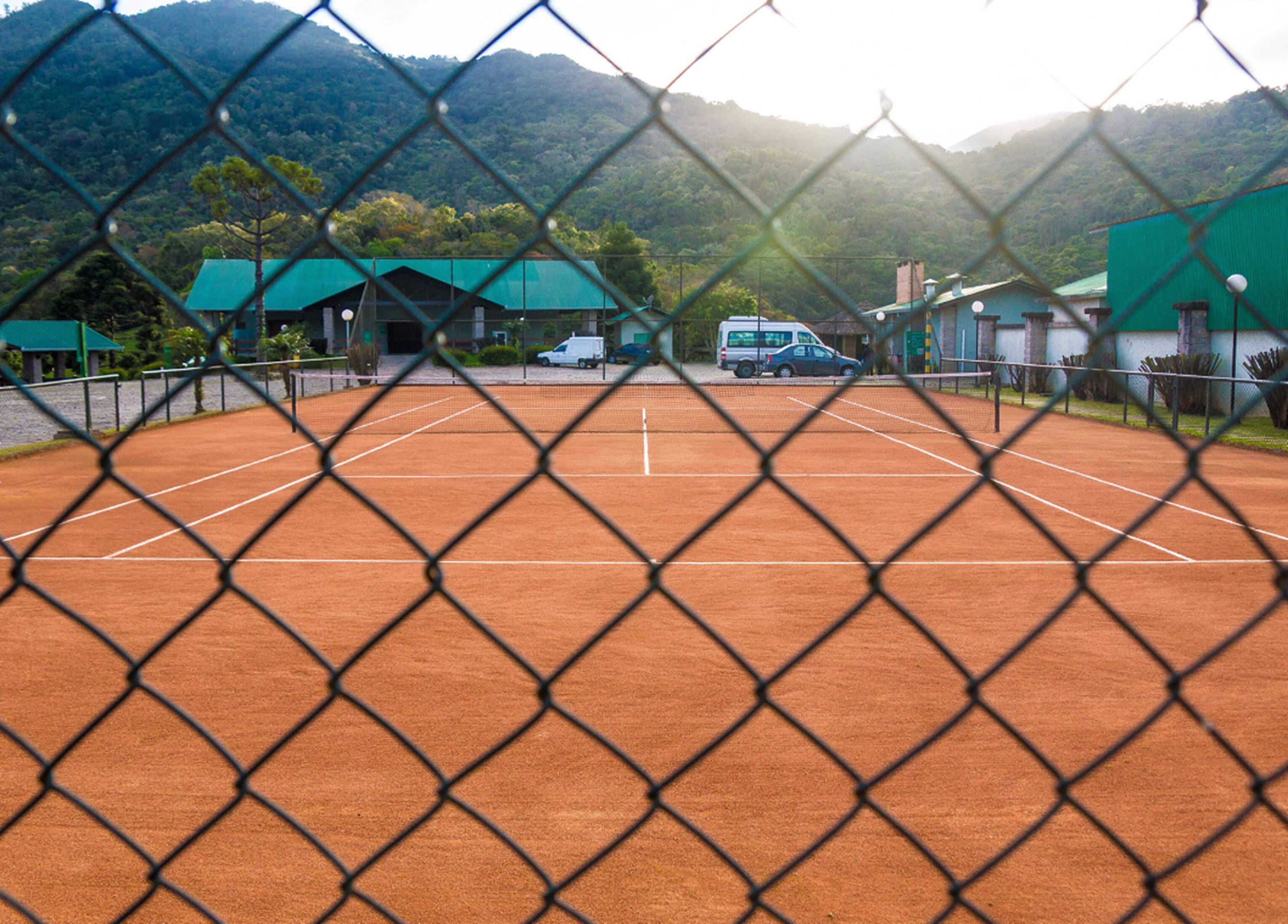Una cancha de tenis rodeada por una cerca de malla metálica, con un edificio de techo verde al fondo.