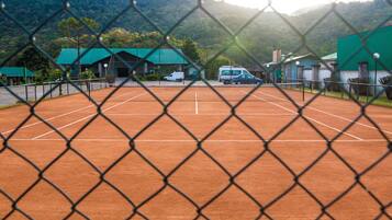 Una cancha de tenis rodeada por una cerca de malla metálica, con un edificio de techo verde al fondo.