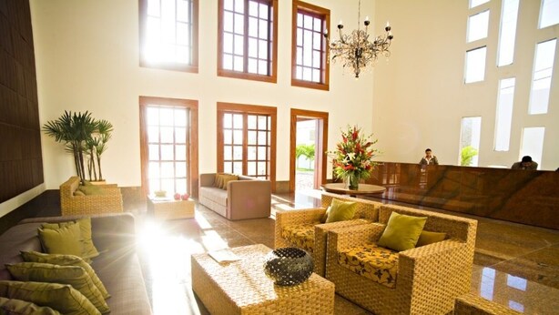 A spacious lobby with a high ceiling, large windows, and a chandelier. There are wicker sofas and a reception desk. - Hotel Villaoeste (Mossoro)