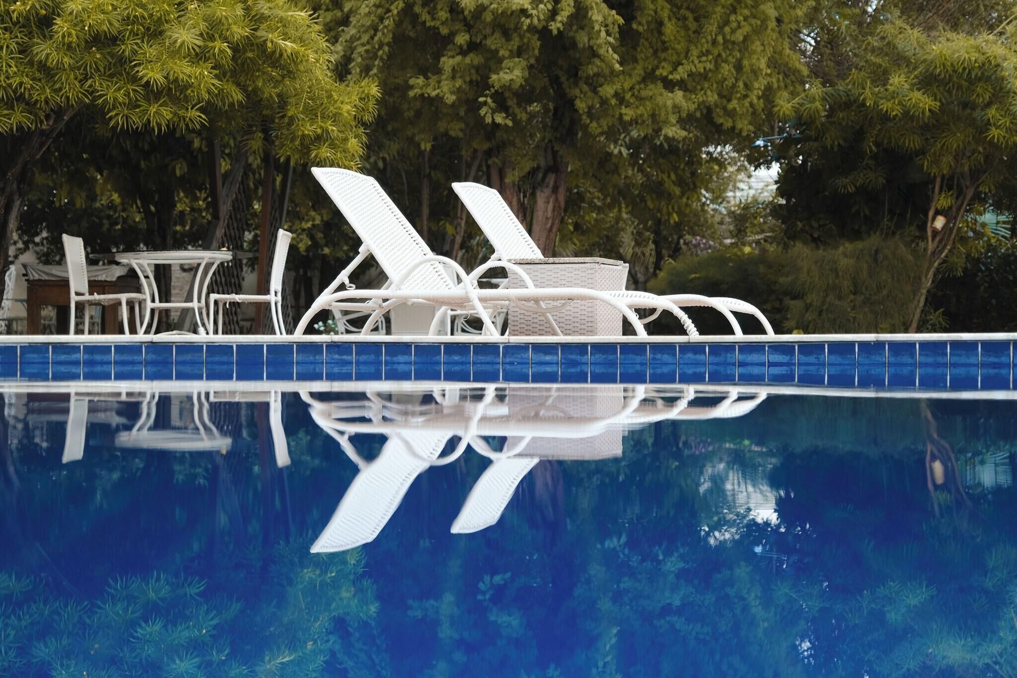Área de piscina com cadeiras de descanso brancas e uma mesa, rodeada por árvores.