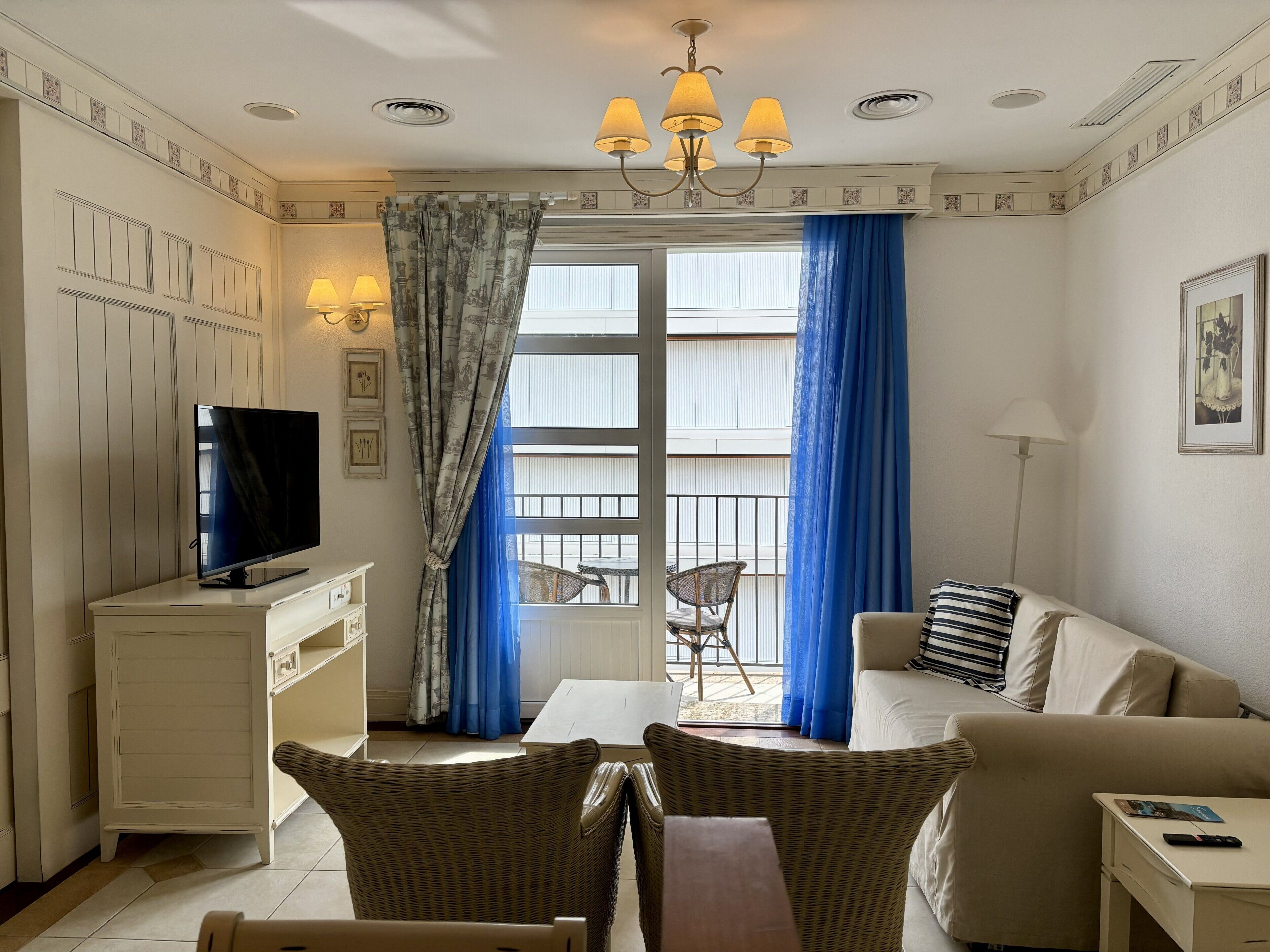 Una sala de estar con televisor, sillas de mimbre, un sofá, una mesita y un balcón con mesa y sillón.