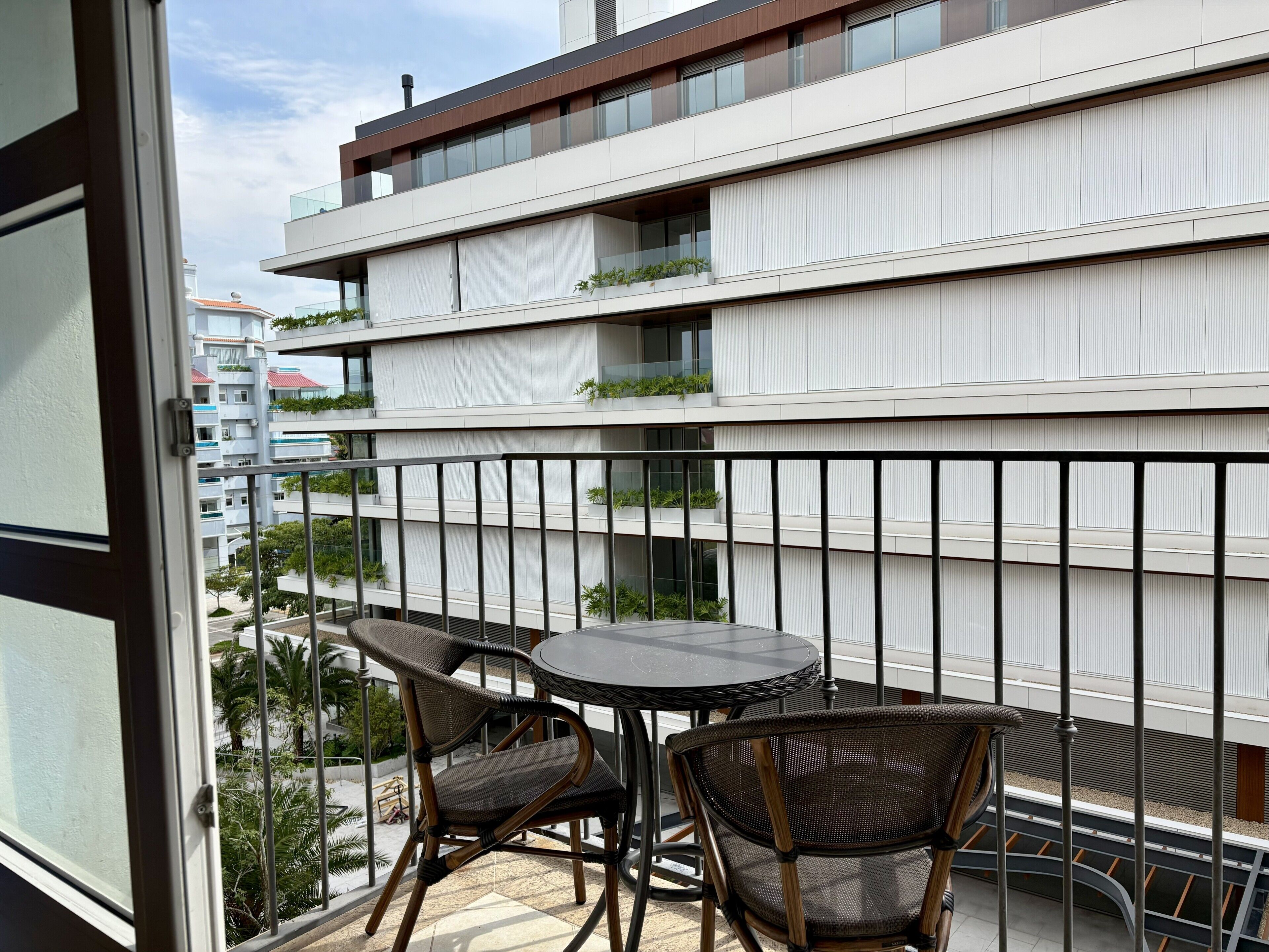 Un balcone con sedie in vimini e un tavolo rotondo, con vista su un edificio moderno con balconi e aree verdi.