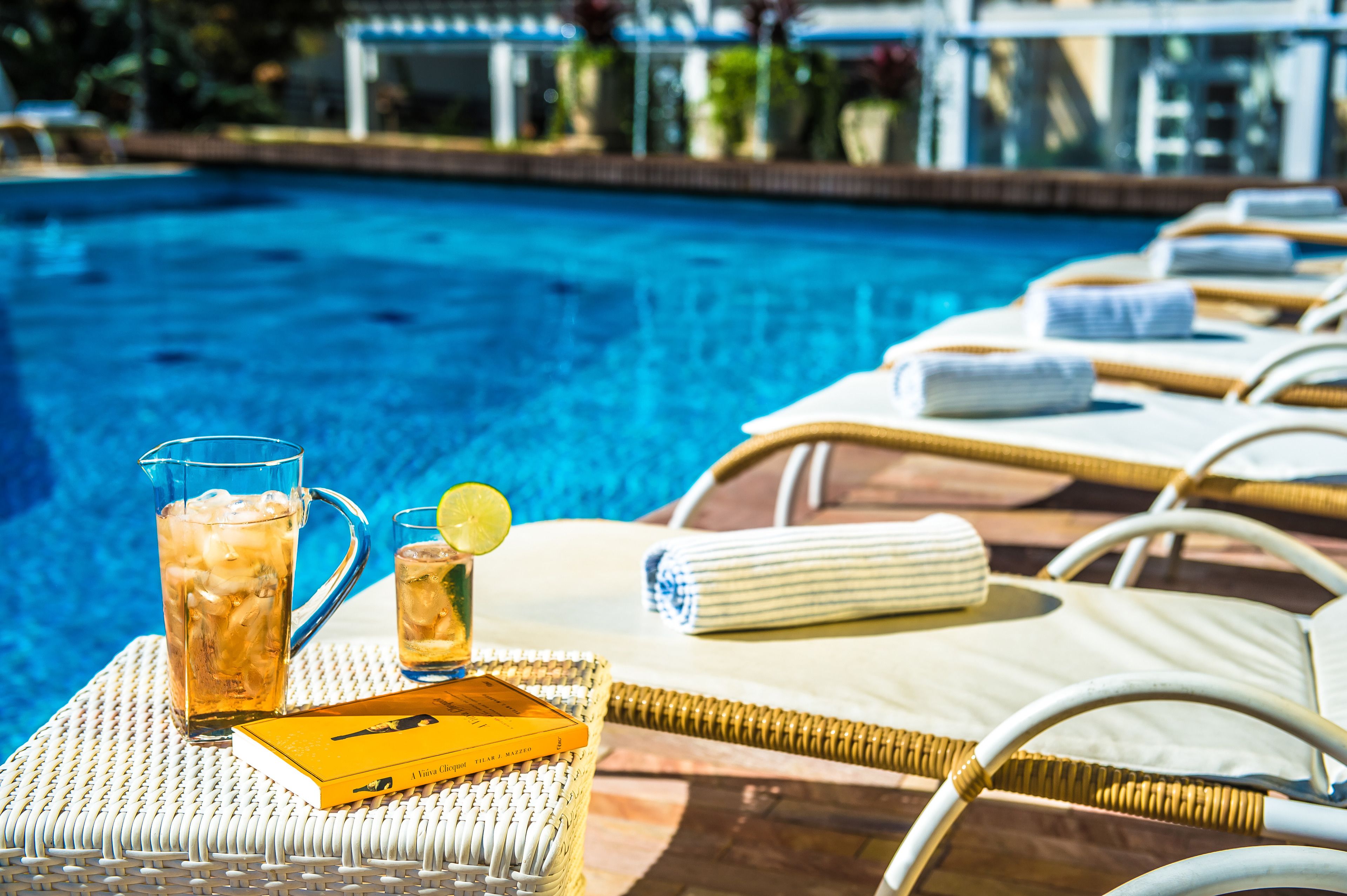 Escena junto a la piscina con sillones de descanso, una mesa con una bebida y un libro, y una vista despejada de la piscina.