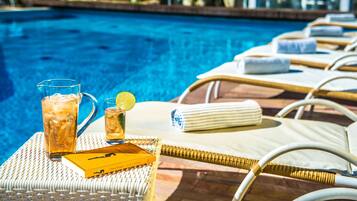 Escena junto a la piscina con sillones de descanso, una mesa con una bebida y un libro, y una vista despejada de la piscina.