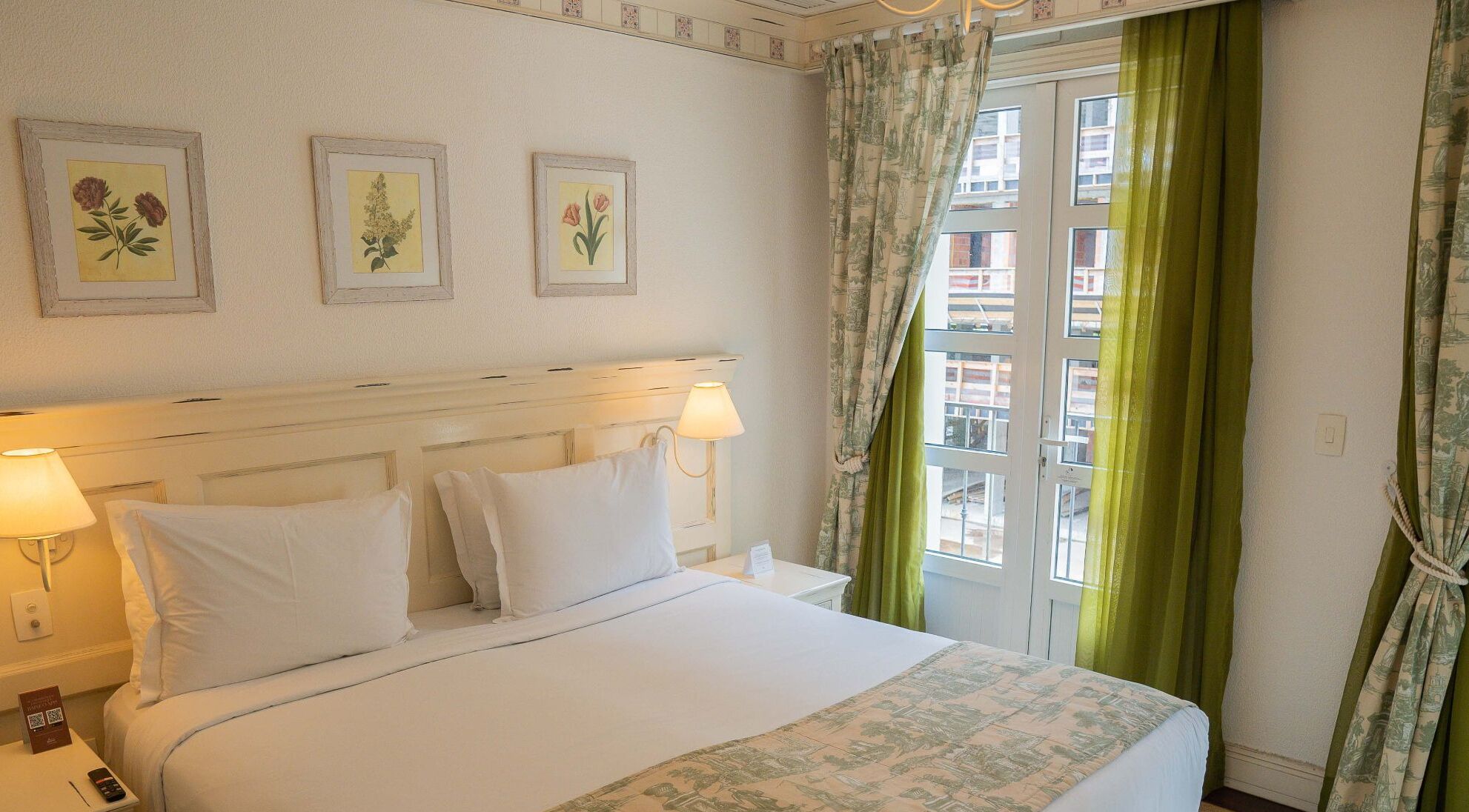 Habitación de hotel con una cama grande, dos lámparas de noche y tres impresiones botánicas enmarcadas en la pared.