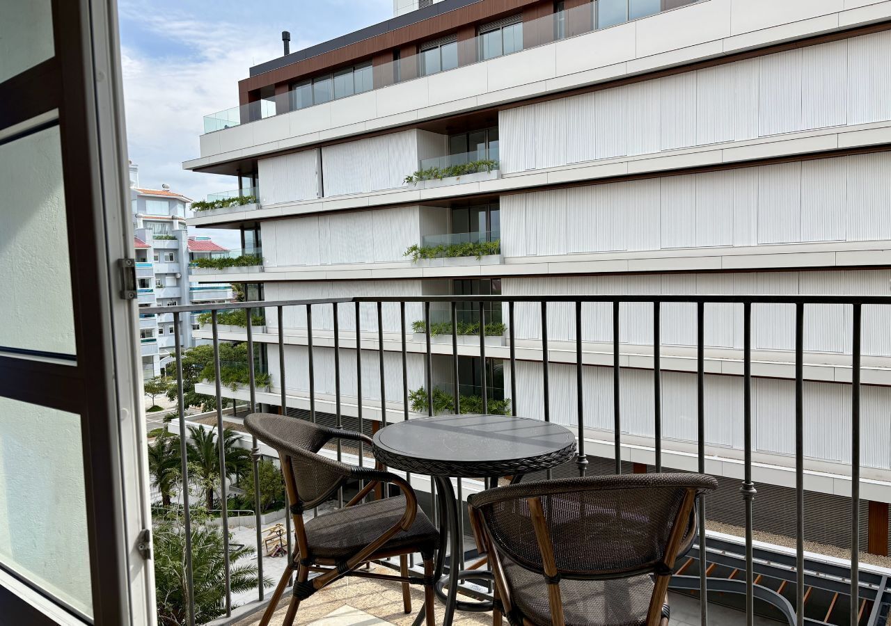 Un balcón con dos sillas de mimbre y una mesita redonda, con vistas a un edificio moderno con balcones y vegetación.