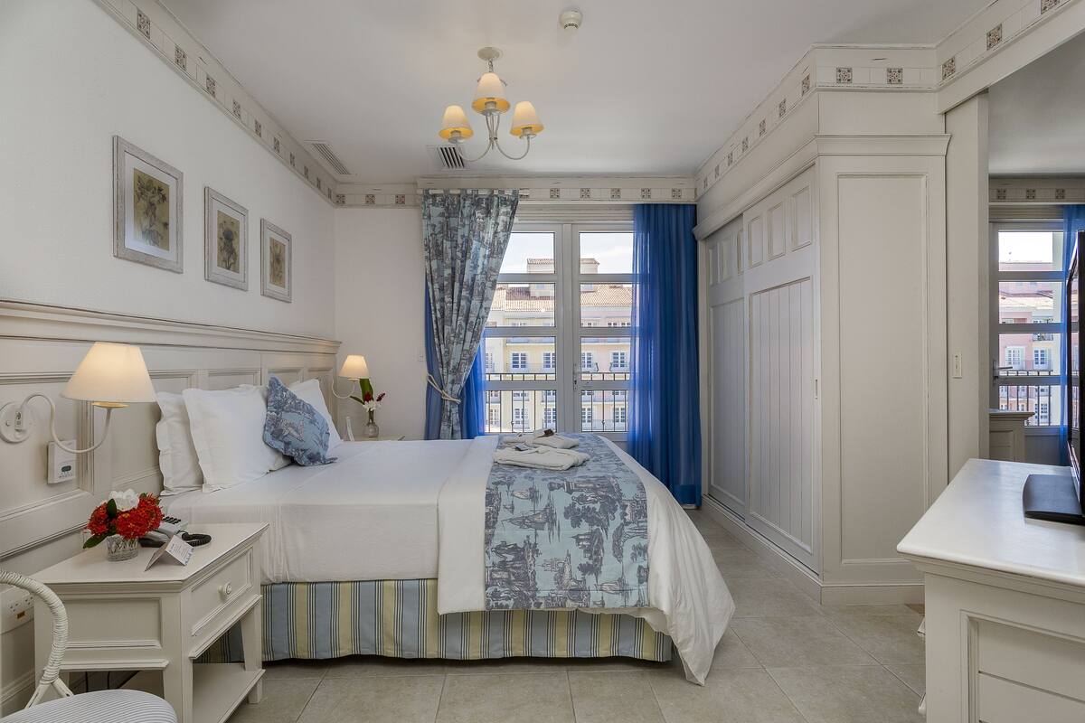 A hotel room with a bed, bedside table, lamp, and a view of buildings through the window.